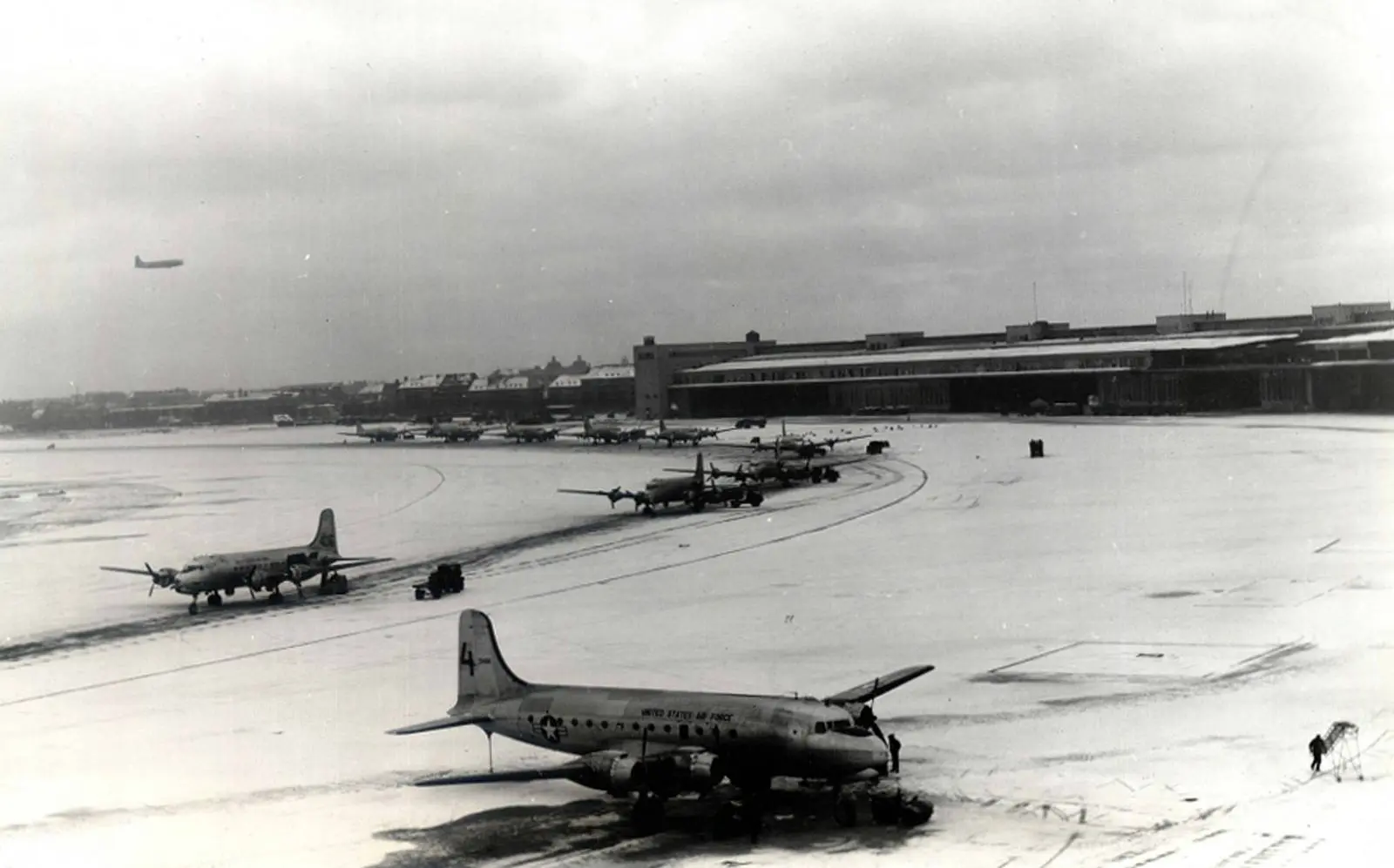 C-54s unload on Tempelhof’s snow-covered ramp while another transport comes in overhead. The airlift delivered supplies to Berlin every single day.