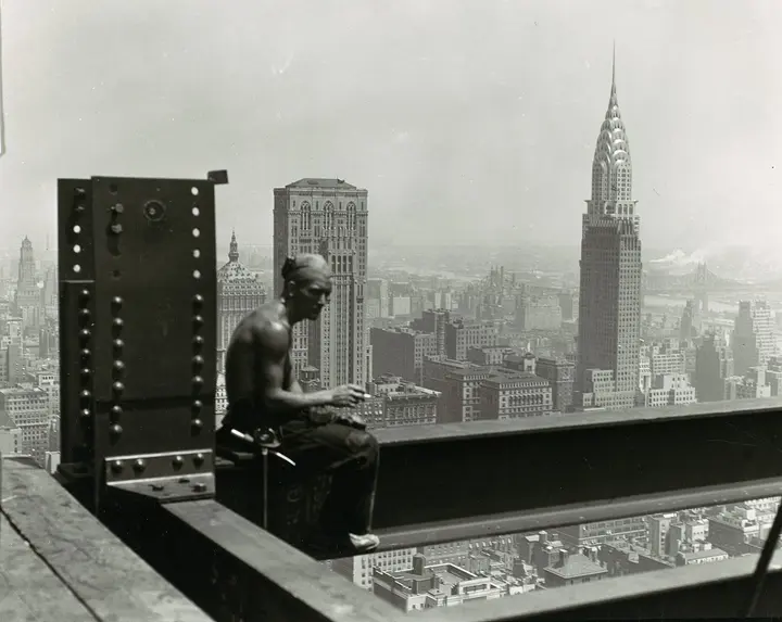 A worker takes a break on a beam hundreds of feet above Manhattan, with the Chrysler Building on the skyline.