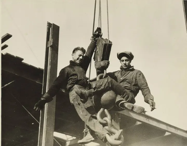 Two young structural workers ride a derrick hook on the rising frame of the Empire State Building.