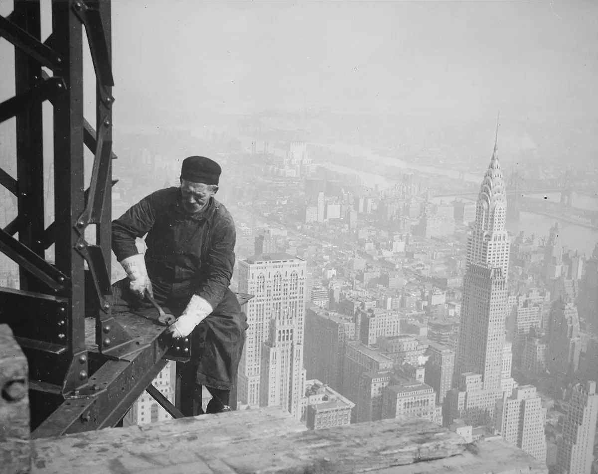 A structural worker bolts a beam high on the Empire State Building, with the Chrysler Building behind him.