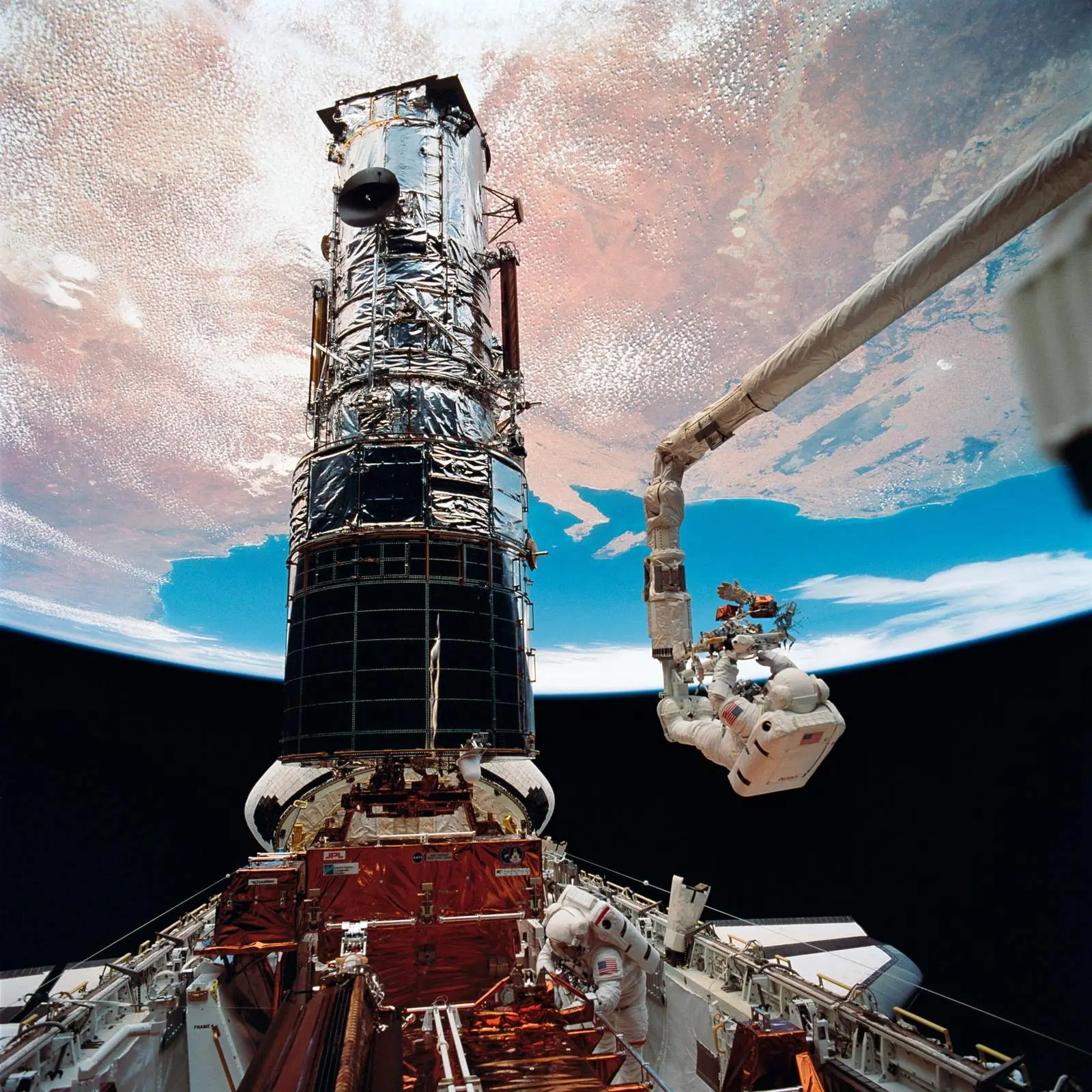 Story Musgrave rides the robot arm toward the top of Hubble on the fifth and final spacewalk, with Hubble standing upright in Endeavour’s payload bay above Earth, 9 December 1993 (STS061-98-050).