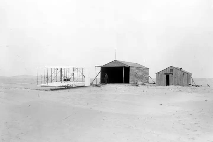 The 1903 Flyer outside the Wrights' hangar and their workshop-living quarters at Kill Devil Hills, N.C., November 24, 1903