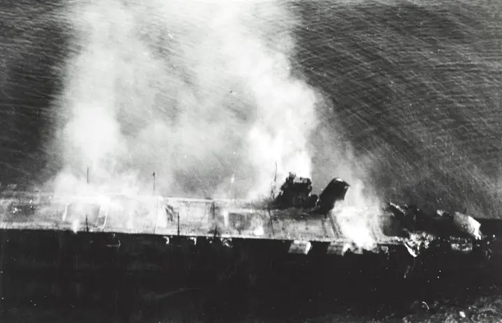The Japanese carrier Hiryu burning at sunrise on 5 June 1942, her forward elevator thrown upright by the explosions in her hangar (NH 73064).