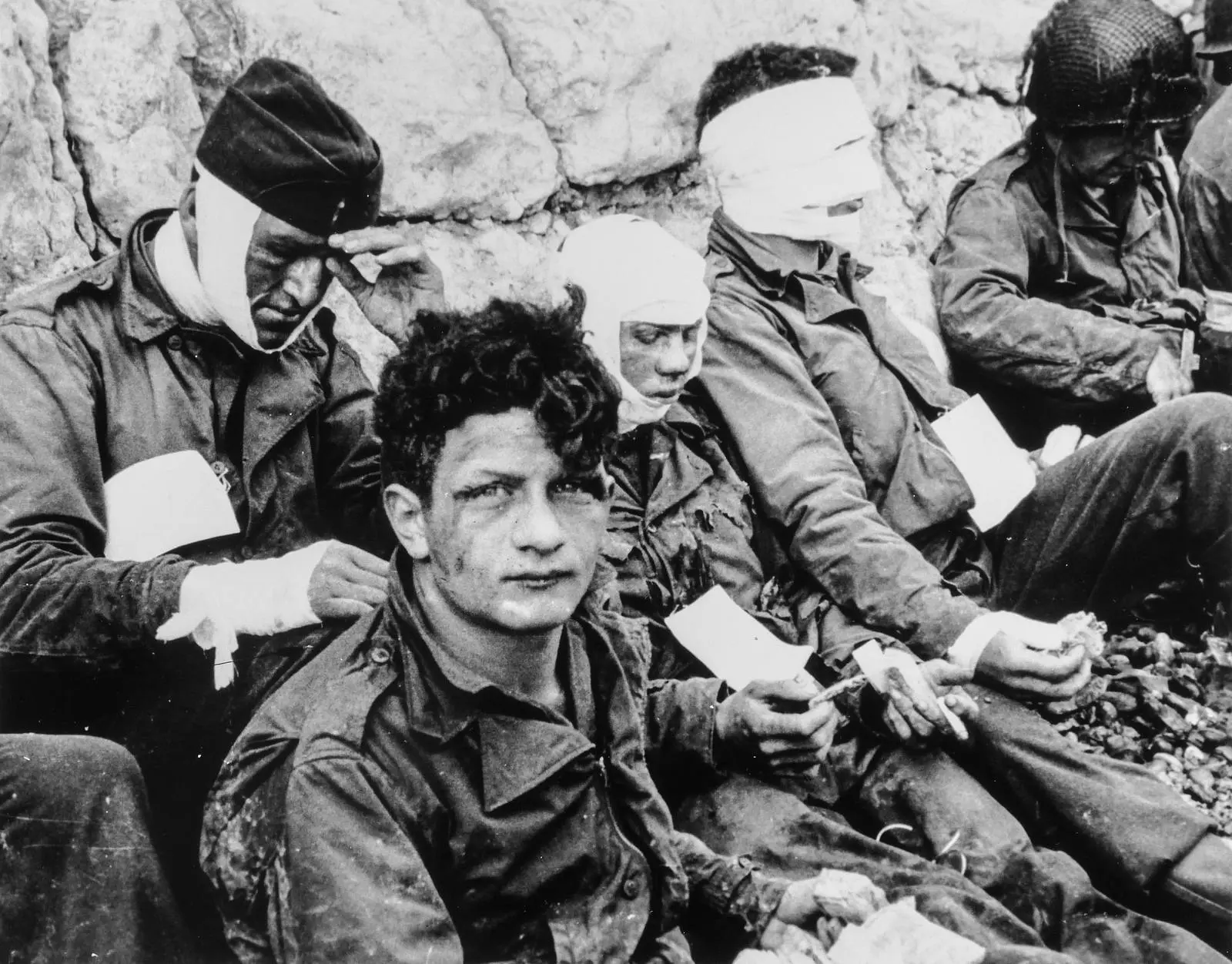 Wounded men of the 16th Infantry Regiment wait at the foot of the chalk cliffs on Omaha Beach for evacuation to a field hospital, 6 June 1944.